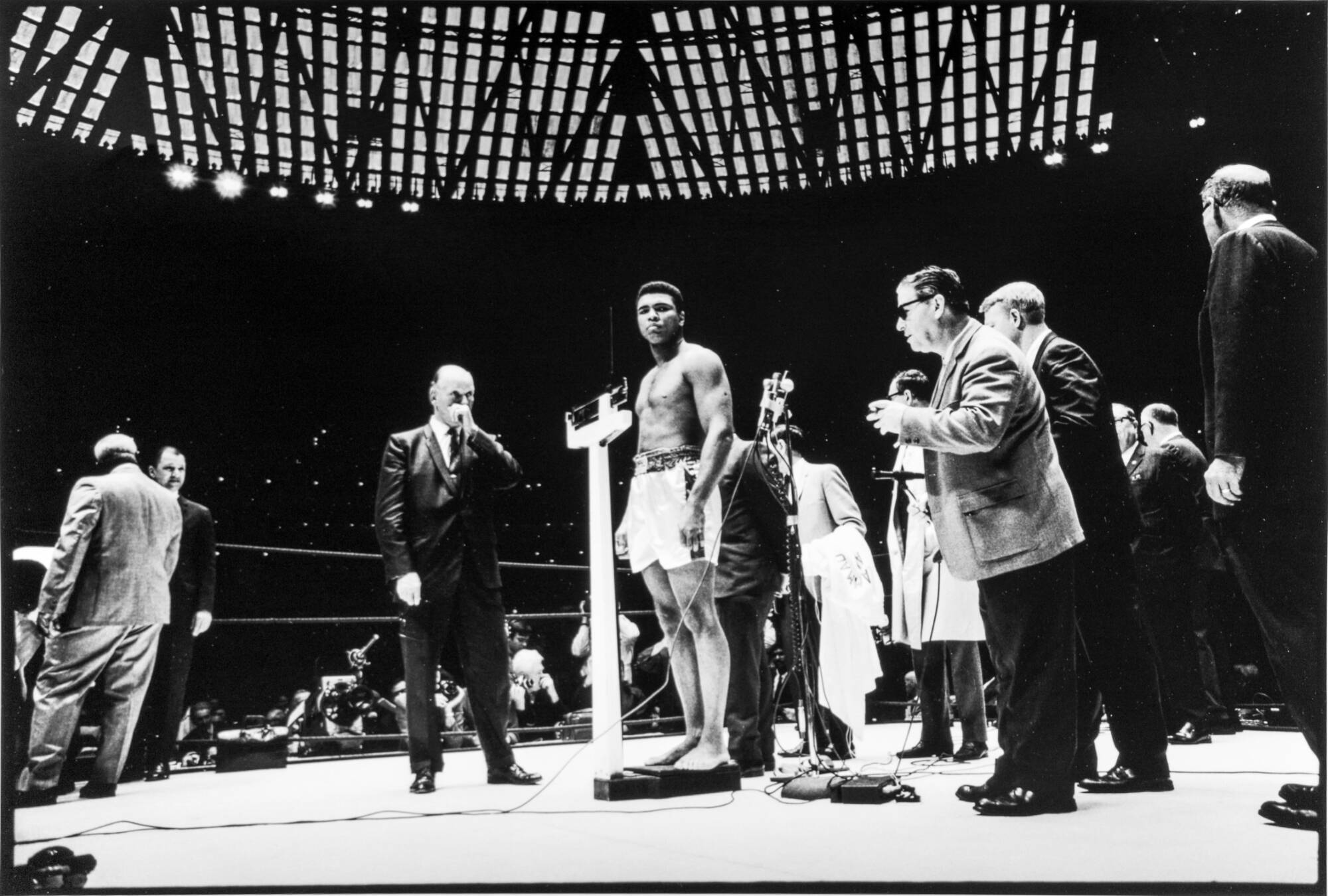 A black and white vertical photograph of Muhammad Ali being weighted in a boxing ring. He is wearing gym shorts